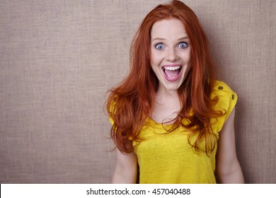Pretty young redhead woman with a happy surprised expression of wide eyed delight standing against a beige background with copy space