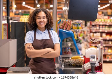 Joven muy sonriente trabajadora de ventas afroamericana con los brazos cruzados por el pecho mirándote en la caja registradora durante el trabajo