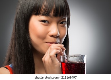 Pretty Young Smiling Dark-haired Asian Woman Drinking Her Favorite Cherry Juice Through The Straw. Isolated On Black Background