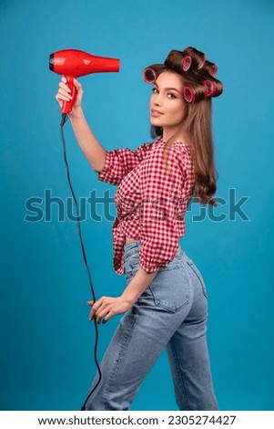 Pretty young smiling girl in big red curlers drying hair with red dryer. Studio work with attractive model in checkered red shirt and jeans posing  on blue background isolated.