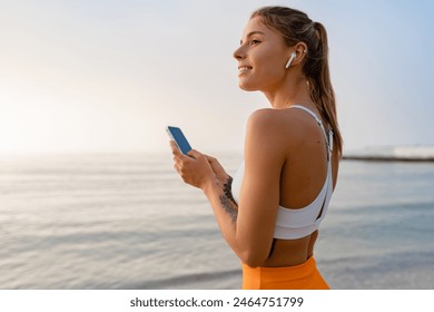 pretty young smiling woman doing sports in the morning by the sea beach in stylish sport outfit sportswear, skinny strong body, healthy fit lifestyle, using smart phone listening to music on earpods - Powered by Shutterstock