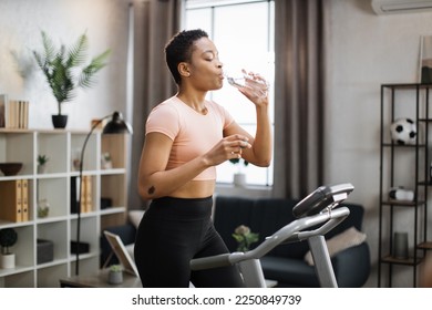 Pretty young sports african woman drinking water from bottle doing fitness exercise, running on treadmill . Athletic and muscular female having workout indoor, at modern apartment at home. - Powered by Shutterstock