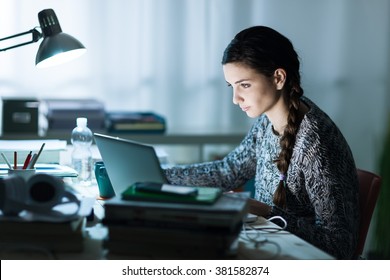 Pretty young student sitting at desk and doing her homework, she is connecting to the internet with a laptop - Powered by Shutterstock