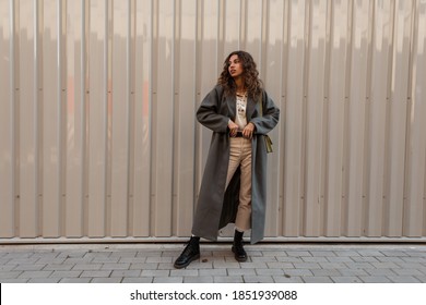 Pretty Young Stylish Woman Model With Curly Hair In A Fashionable Long Coat With Boots Straightens Her Belt Near A Metal Wall. Female Casual Fashion And Style