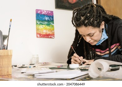 Pretty Young White Girl, Hispanic Latin Art Director, With Dreadlocks, In Her Workshop Working, On Her Table, Gluing With Precision Paper And Styrofoam With A Fine Brush And White Glue.