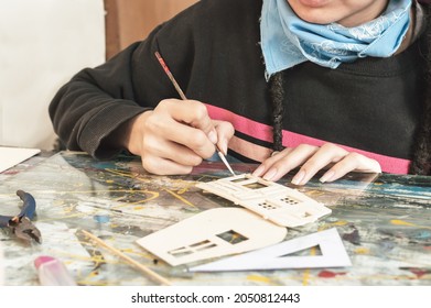 Pretty Young White Girl Hispanic Latina Art Director With Dreadlocks And A Blue Bandanna, Painting Small Parts Of A Mockup With A Brush On Her Work Table In Her Artistic Design Studio.