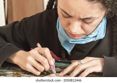 Pretty Young White Girl Hispanic Latina Art Director, With Dreadlocks And A Frown On Her Face, Sitting At Her Work Table Cutting Cardboard In Great Detail With A Cutter, In Her Workshop.