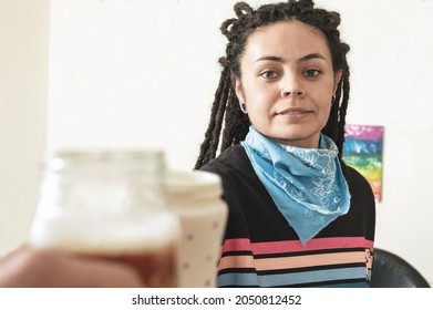 Pretty Young White Girl Hispanic Latina Art Director, With Dreadlocks, Black Sweater And Blue Bandanna, In Her Art Studio, Smiling Making A Toast With A Glass Of Beer, While Resting.