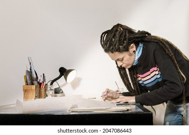 Pretty Young White Hispanic Latin Art Director Girl With Dreadlocks, In Her Studio With A Cutter In Hand Working Cutting Styrofoam, With Papers And Work Implements On A Wooden Table.