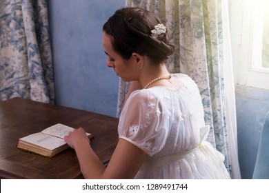 Pretty young woman in authentic regency dress reading a book in vintage room