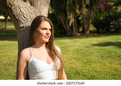 Pretty Young Woman, Blonde, Blue Eyes, Leaning Against A Tree Trunk In An Outdoor Park On A Sunny Day. Concept Beauty, Fashion, Model, Nature, Spring.