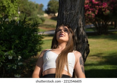 Pretty Young Woman, Blonde, Blue Eyes, Leaning Against A Tree Trunk In An Outdoor Park On A Sunny Day. Concept Beauty, Fashion, Model, Nature, Spring.