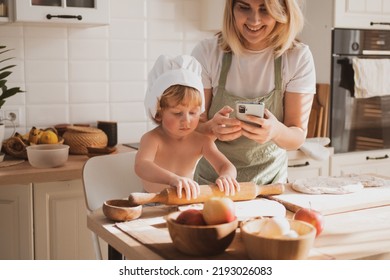 Pretty Young Woman In Chef's Apron And Her Little Son Are Preparing Apple Pie At Home In Cozy Kitchen. Kid In Cute Chef's Hat. She Takes Pictures Of The Process Of Making Cake