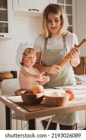 Pretty Young Woman In Chef's Apron And Her Little Son Are Preparing Apple Pie At Home In Cozy Kitchen. Kid In Cute Chef's Hat