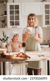 Pretty Young Woman In Chef's Apron And Her Little Son Are Preparing Apple Pie At Home In Cozy Kitchen. Kid In Cute Chef's Hat