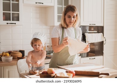 Pretty Young Woman In Chef's Apron And Her Little Son Are Preparing Apple Pie At Home In Cozy Kitchen. Kid In Cute Chef's Hat