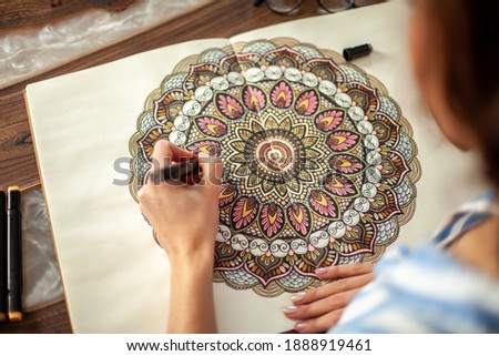 Pretty young woman coloring the mandala with colored markers. An album with a circular pattern lies on the table. Hobby and relaxation concept.