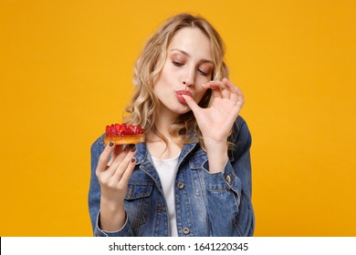 Pretty Young Woman In Denim Clothes Isolated On Yellow Orange Background. Proper Nutrition Or Sweets, Dessert Fast Food, Dieting Concept. Mock Up Copy Space. Holding Strawberry Cake, Licking Fingers