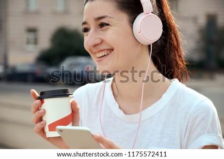 Pretty young woman drinking takeout coffee cup, using smart phone, listening to music in headphones outdoors. Urban lifestyle concept. Healthy, positive, cheerful morning running.