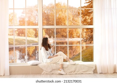 Pretty young woman drinking tea while sitting near window on autumn day - Powered by Shutterstock