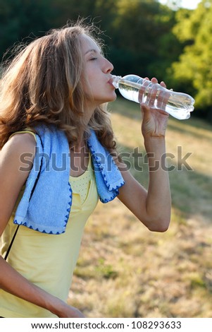 Pretty young woman drinking water after fitness workout against the background of nature on a sunny day