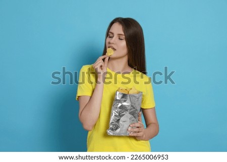 Pretty young woman eating tasty potato chips on light blue background