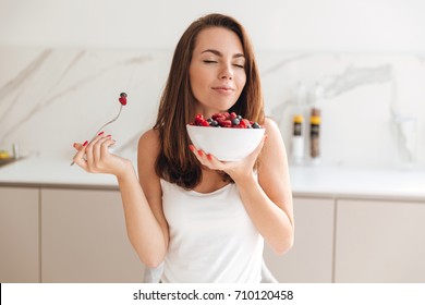 Pretty young woman enjoys eating fresh berries from a bowl while sitting in a kitchen