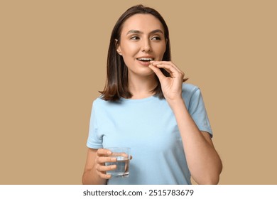 Pretty young woman with glass of water taking pill on brown background - Powered by Shutterstock