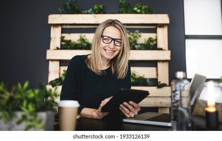 Pretty Young Woman With Glasses Sits In A Modern, Sustainable Office With Lots Of Green Ecological Plants And Works On Her Tablet And Is Happy
