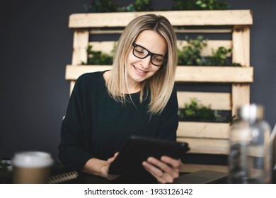 Pretty Young Woman With Glasses Sits In A Modern, Sustainable Office With Lots Of Green Ecological Plants And Works On Her Tablet And Is Happy
