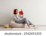 Pretty young woman in headphones reading book on beanbag chair at home