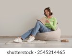 Pretty young woman in headphones reading book on beanbag chair at home