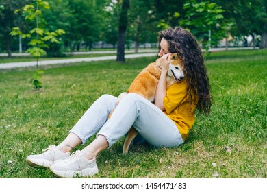 Pretty young woman is hugging shiba inu dog and smiling sitting on grass in park enjoying friendship with animal. People, affection and cute pets concept.