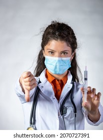 Pretty Young Woman In Lab Coat Wearing Protective Mask, Holding Syringe With Vaccine And Showing Thumb Down