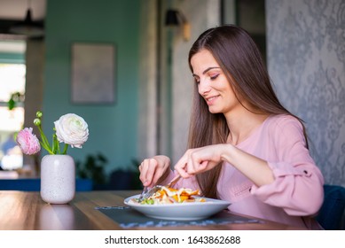 Pretty Young Woman With Long Hair And In Pink Shirt Eating A Salad At The Nice Restaurant. Eating Alone. Comfortable, Happy, Smiling, Carefree.