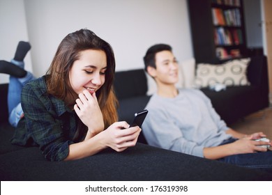 Pretty Young Woman Lying On Couch Texting On Mobile Phone While Young Man Playing Video Game In Background At Home. Mixed Race Teenage Couple In Living Room Indoors.