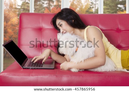 Pretty young woman lying on sofa while using laptop computer with her dog, shot with autumn background on the window