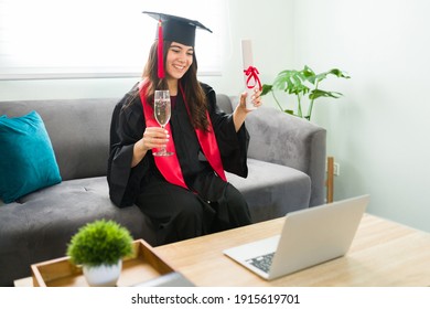 Pretty Young Woman Making A Toast With A Glass Of Wine During Her Virtual Graduation Ceremony. Female Graduate Celebrating Her Academic Success In A Video Call Because Of The Coronavirus Pandemic