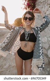 Pretty Young Woman, Model Having Fun On Vacation, Great Summer Time. Wearing Stylish Sunglasses, Black Fashionable Bikini, Cardigan, Cape With Ornaments. Posing Near Rock On Beach.