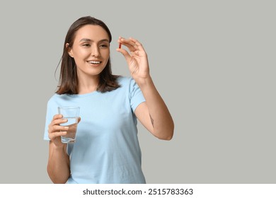 Mujer joven bonita con la píldora y el vaso de agua sobre fondo gris