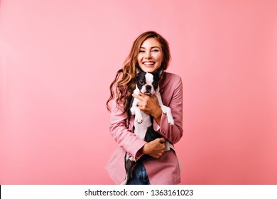 Pretty young woman in pink jacket playing with her dog. Indoor photo of european jocund girl posing with french bulldog.