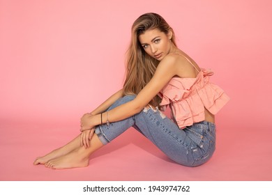 Pretty Young Woman In Pink Shirt And Jeans Sitting On Floor On Pink Background In Studio Barefoot, Copy Space