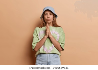 Pretty young woman pleading and asking for something with clasped hands has begging facial expression makes request dressed in casual clothing isolatd over brown studio background. Please help me