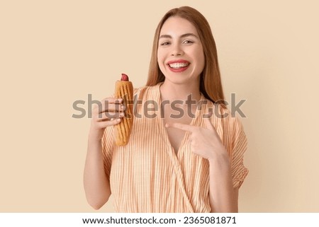 Pretty young woman pointing at tasty french hot dog on beige background