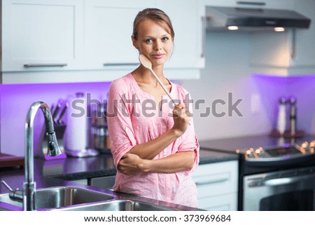 Pretty, young woman posing in her modern kitchen, holding a wooden spoon, ready to cook something delicious (shallow DOF; color toned image)