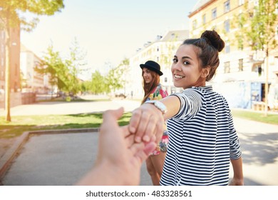 Pretty Young Woman Pulling A Man Along By The Hand Turning To Look Back At Him With A Happy Smile As Her Friend Waits In The Background