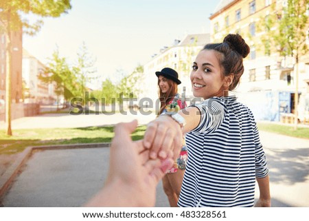 Pretty young woman pulling a man along by the hand turning to look back at him with a happy smile as her friend waits in the background