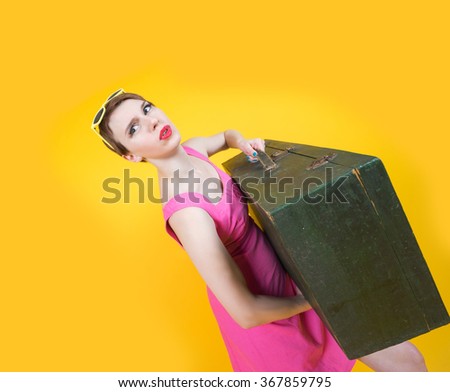 Pretty young woman in red dress holding heavy suitcase and shocked. isolated on yellow background. wide angle shoot.