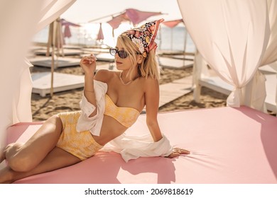 Pretty young woman relaxes lying on beach bed with soft pink mattress. Beauty with light short hair in yellow two-piece swimsuit wearing sunglasses. Summer vacation concept on photo. - Powered by Shutterstock