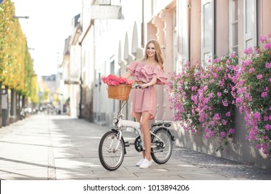 Pretty young woman is riding on a white bicycle in the city. She is smiling and wearing a pink off shoulder dress. On her bike is basket with pink roses.